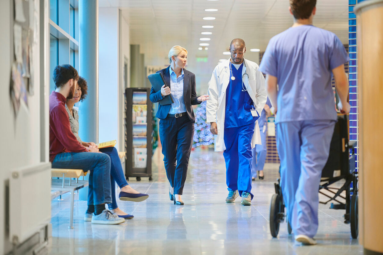 un médecin de sexe masculin d'âge moyen parle avec une femme d'affaires, une vendeuse dans le couloir de l'hôpital alors qu'ils se rendent à une réunion, avec une jeune infirmière et des patients dans le couloir très fréquenté.