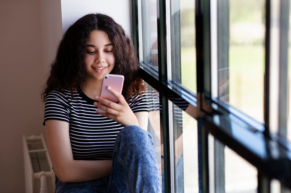 Adolescente lisant un message texte sur un t&eacute;l&eacute;phone portable par une fen&ecirc;tre en verre