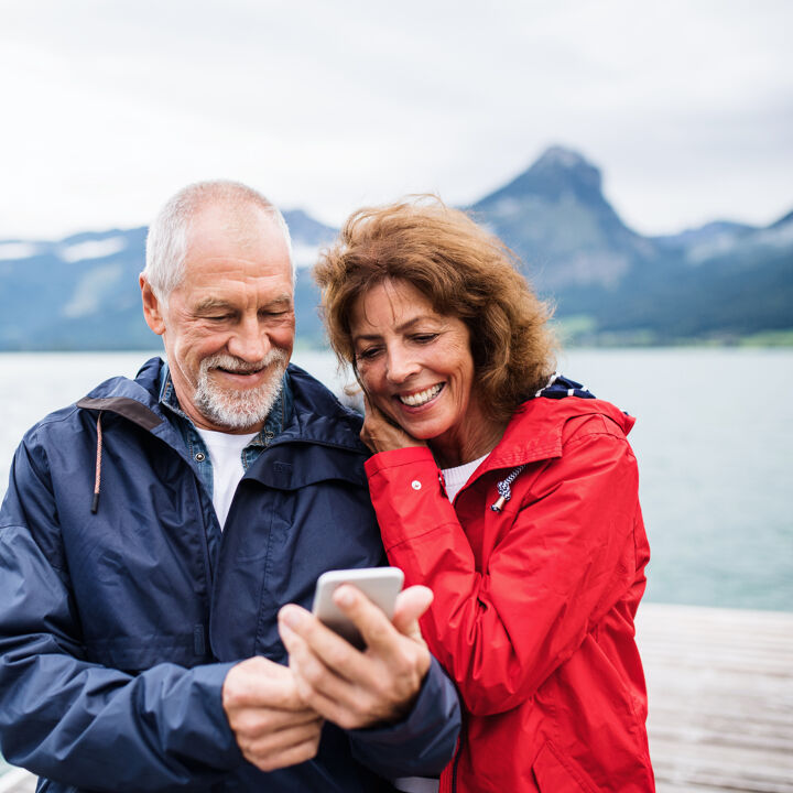 Joyeux couple de personnes âgées debout au bord du lac dans la nature en vacances, à l'aide d'un smartphone.