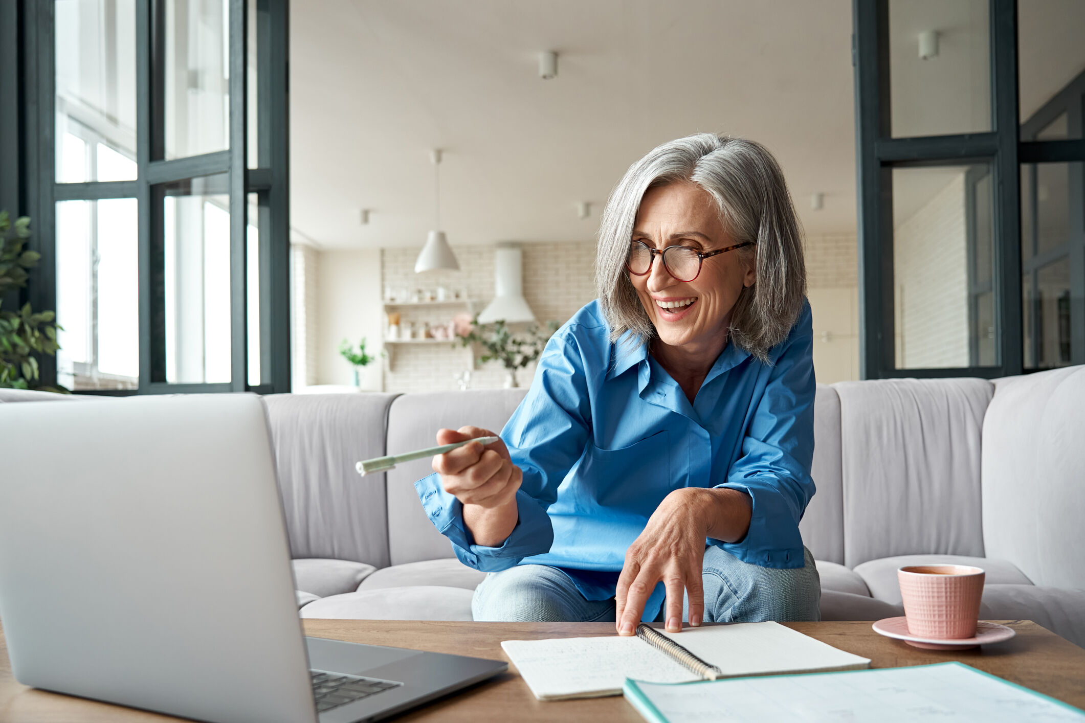 Femme avec des lunettes et une chemise bleu sur son canap&eacute; pointant avec un stylo son ordinateur